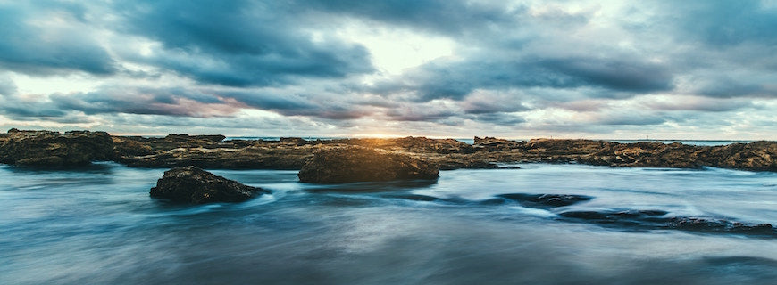 rock-strewn area of ocean, with clouds parting overhead and sun shining down